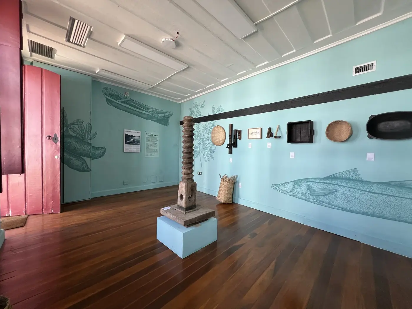 Sala da exposição do Glossário Caiçara de Paraty, com monjolo de rosca e pinturas de peixes na parede